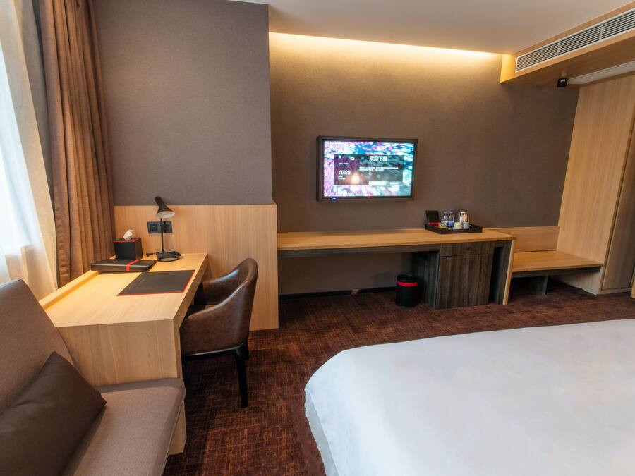 A hotel room with a bed, a desk with a chair, a television mounted on the wall, and a trash bin.
