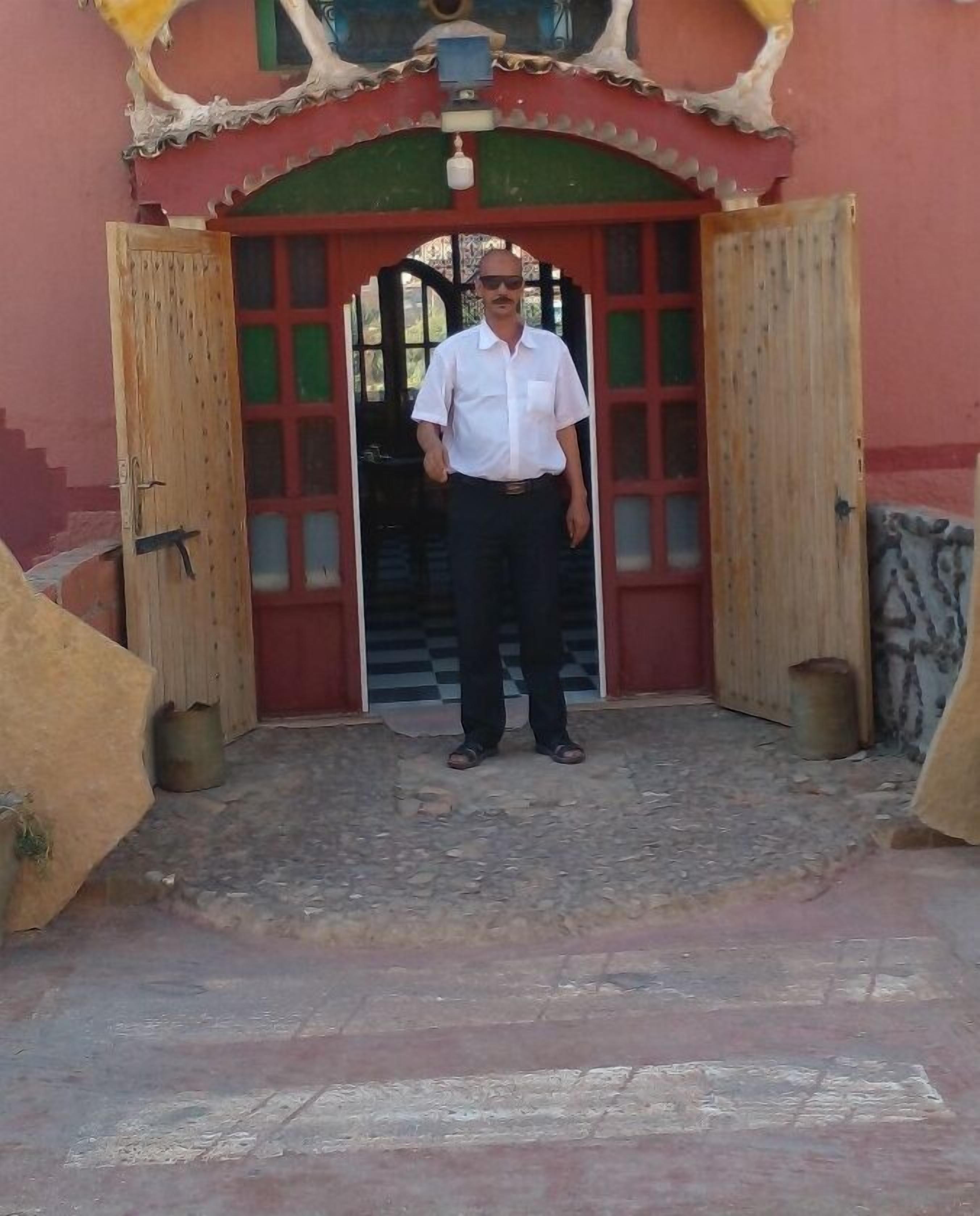 A man standing in front of a building with a red and green door, wooden pillars, and a stone floor.