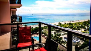 A balcony with red chairs, a pool, and a view of the beach and ocean.