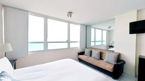 A hotel room with a large bed, a sofa with striped pillows, a TV mounted on the wall, and a window offering a view of the sea. - Castle 1033  Ocean view studio, Pool and Beach Access, FREE Parking, Tennis and more! (Miami Beach)