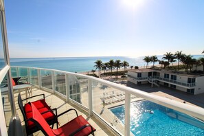 A balcony with red chairs, a pool, and a view of the ocean and palm trees. - Castle M16 Ocean/Pool view, balcony, gym. tennis, & free valet parking! (Miami Beach)