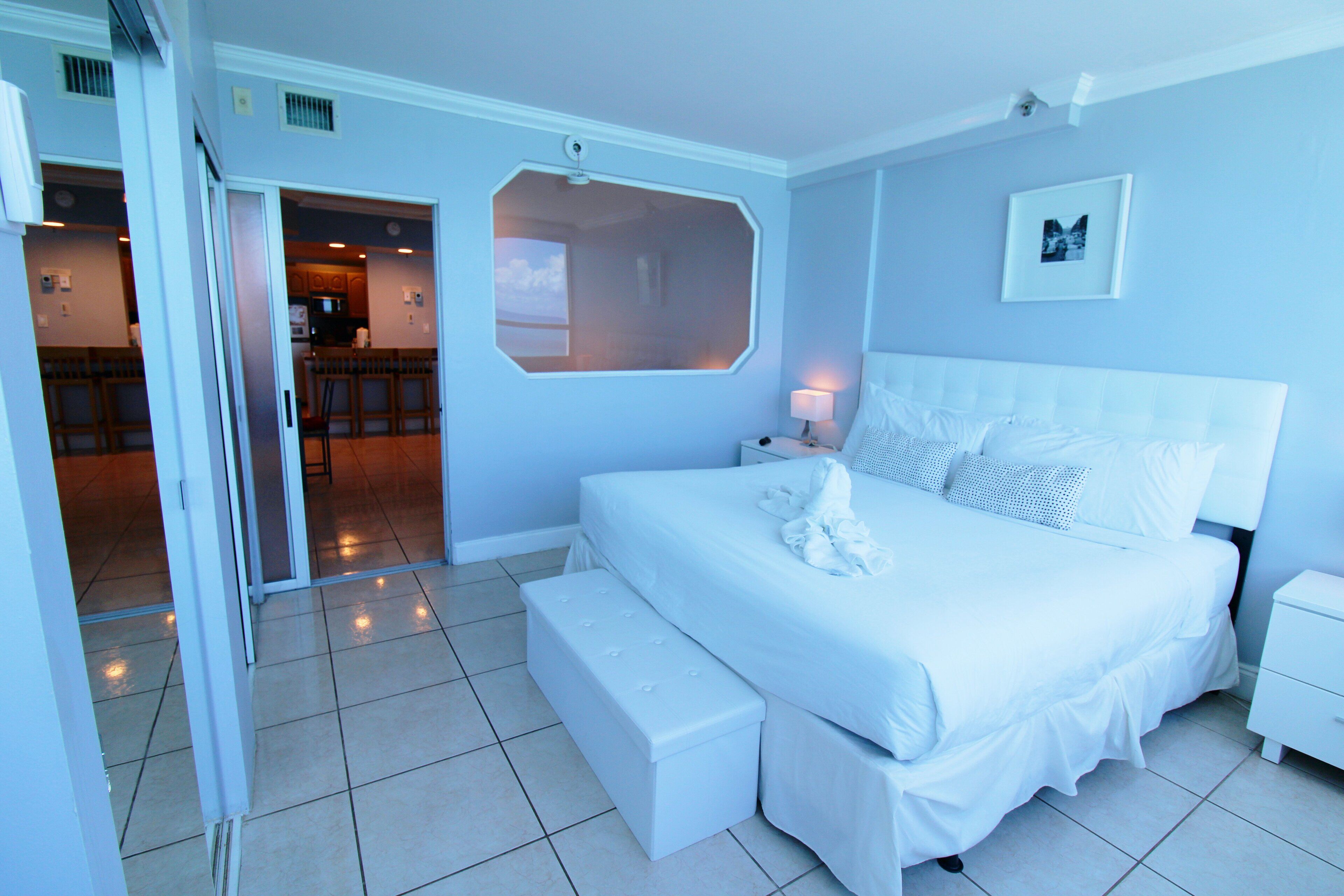 A bedroom with a large bed, a bench, a nightstand, and a view of the kitchen through a window.