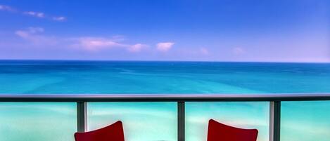 A balcony with a view of the ocean, a round table with a patterned tablecloth, and two red chairs.