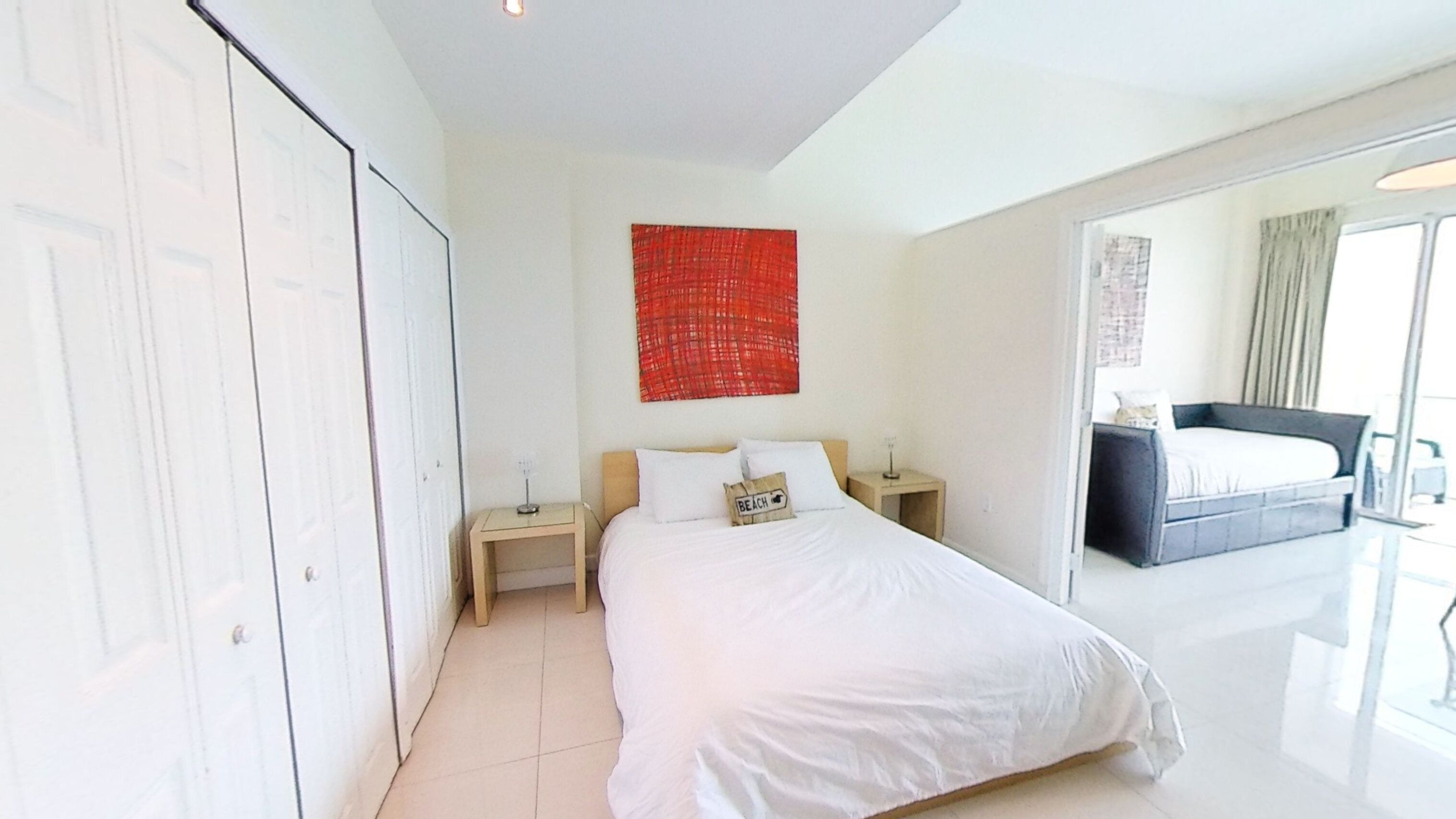 A modern hotel room with a large bed, a bedside table, a red abstract painting, and a view of another room with a bed.