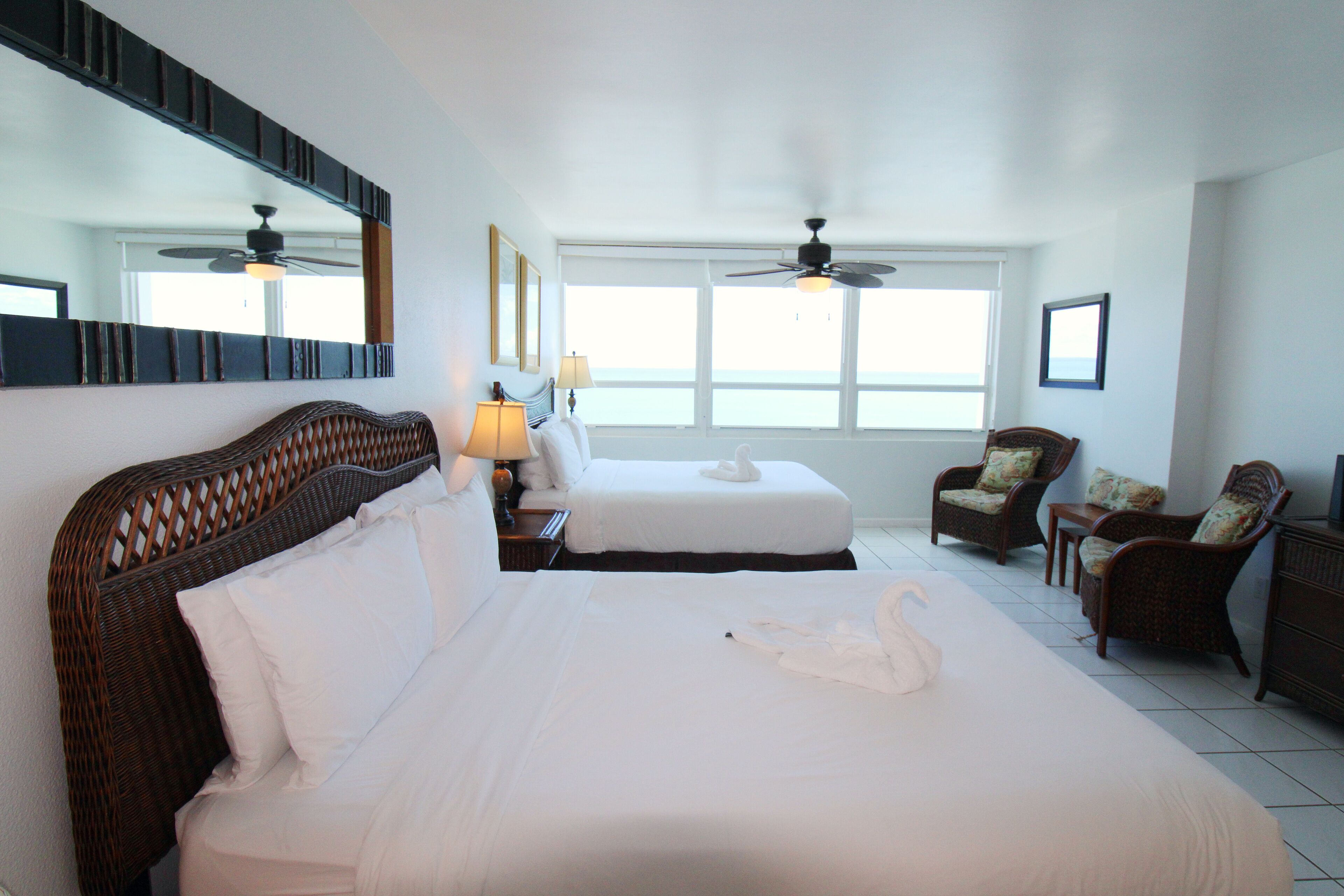 A hotel room with two beds, a large mirror, ceiling fans, wicker furniture, and a view of the sea through large windows.