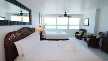 A hotel room with two beds, a large mirror, ceiling fans, wicker furniture, and a view of the sea through large windows.