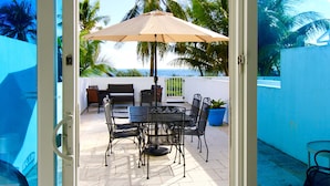 An outdoor patio with a table and chairs, a beige umbrella, and a view of palm trees and the ocean.