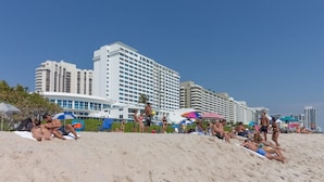 A beach with people sunbathing and swimming, and a large hotel with multiple floors and balconies in the background.