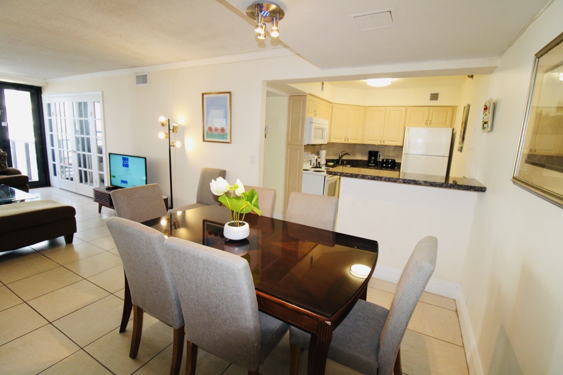 A dining area with a wooden table, grey upholstered chairs, and a vase with white flowers. A television is visible in the background.