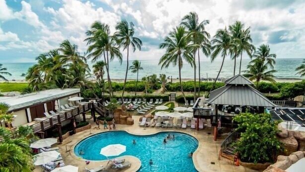 A pool area with lounge chairs and umbrellas, surrounded by palm trees and a view of the beach. - Alexander 1211 with Beach Access, 2 pools, sauna (Miami Beach)