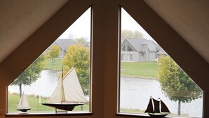 Uitzicht vanuit een driehoekig raam op een rustig meer met zeilboten, een gazon en een gebouw op de achtergrond.
