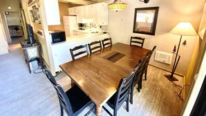 A dining area with a wooden table and chairs, a kitchen in the background, and a hanging light fixture. - Mountain Retreat.                  (Mammoth Lakes)