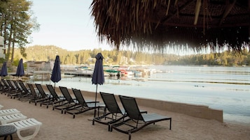 Un área de descanso junto a la playa con tumbonas y sombrillas, con vista a un lago tranquilo con barcos.