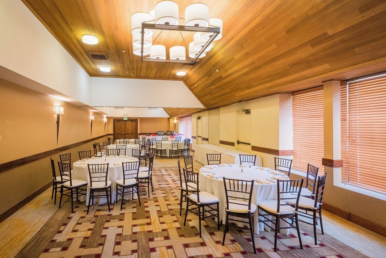 Un salón de banquetes con mesas redondas y sillas, techo de madera y apliques en las paredes.