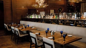 Interior de restaurante con mesas de madera, bancos acolchados blancos y una zona de bar con diferentes botellas.