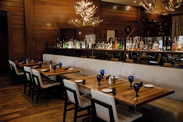 Interior de restaurante con mesas de madera, bancos acolchados blancos y una zona de bar con diferentes botellas.