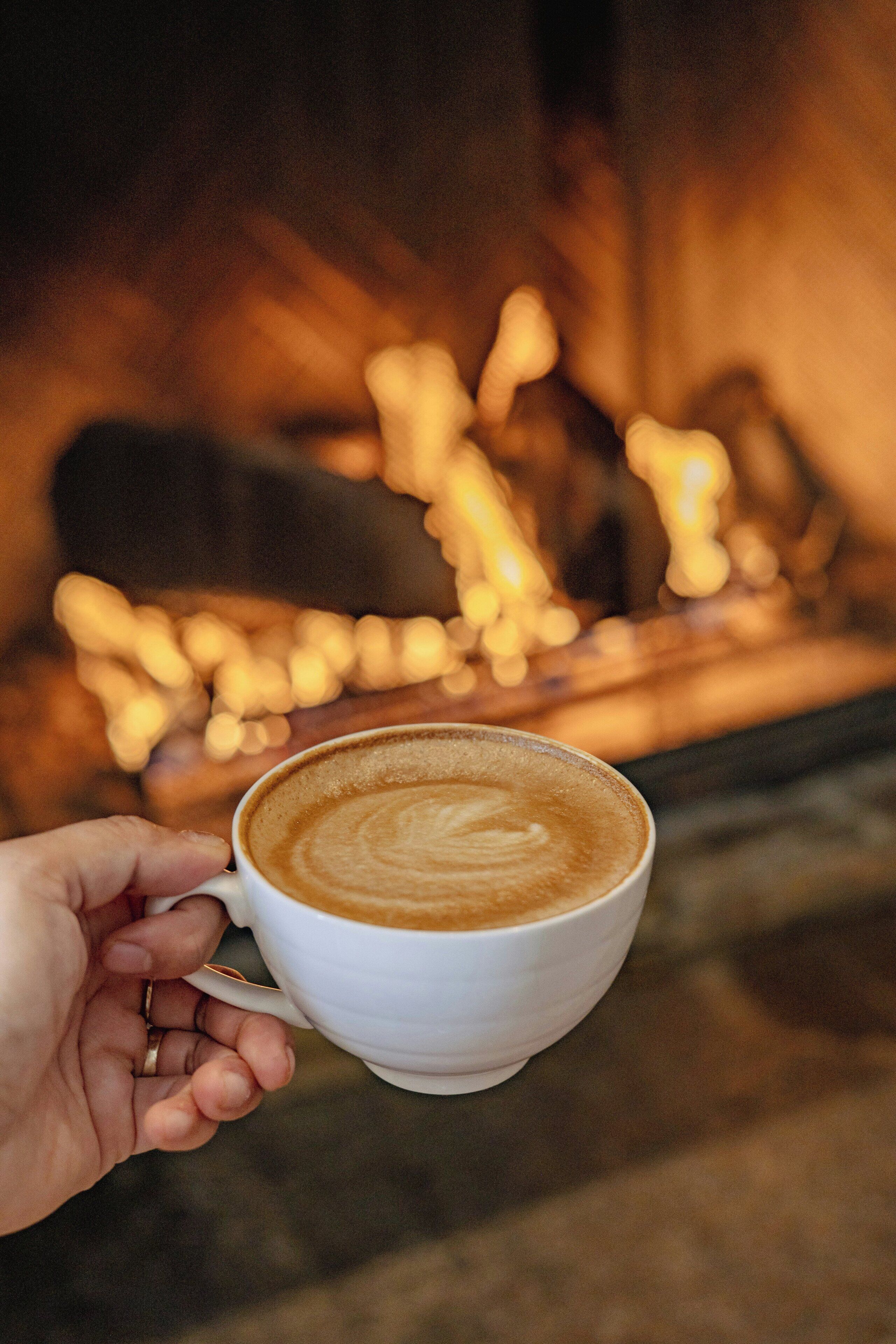 A hand holding a cup of coffee with a latte art design, in front of a fireplace with visible flames.