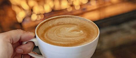 A hand holding a cup of coffee with a latte art design, in front of a fireplace with visible flames.