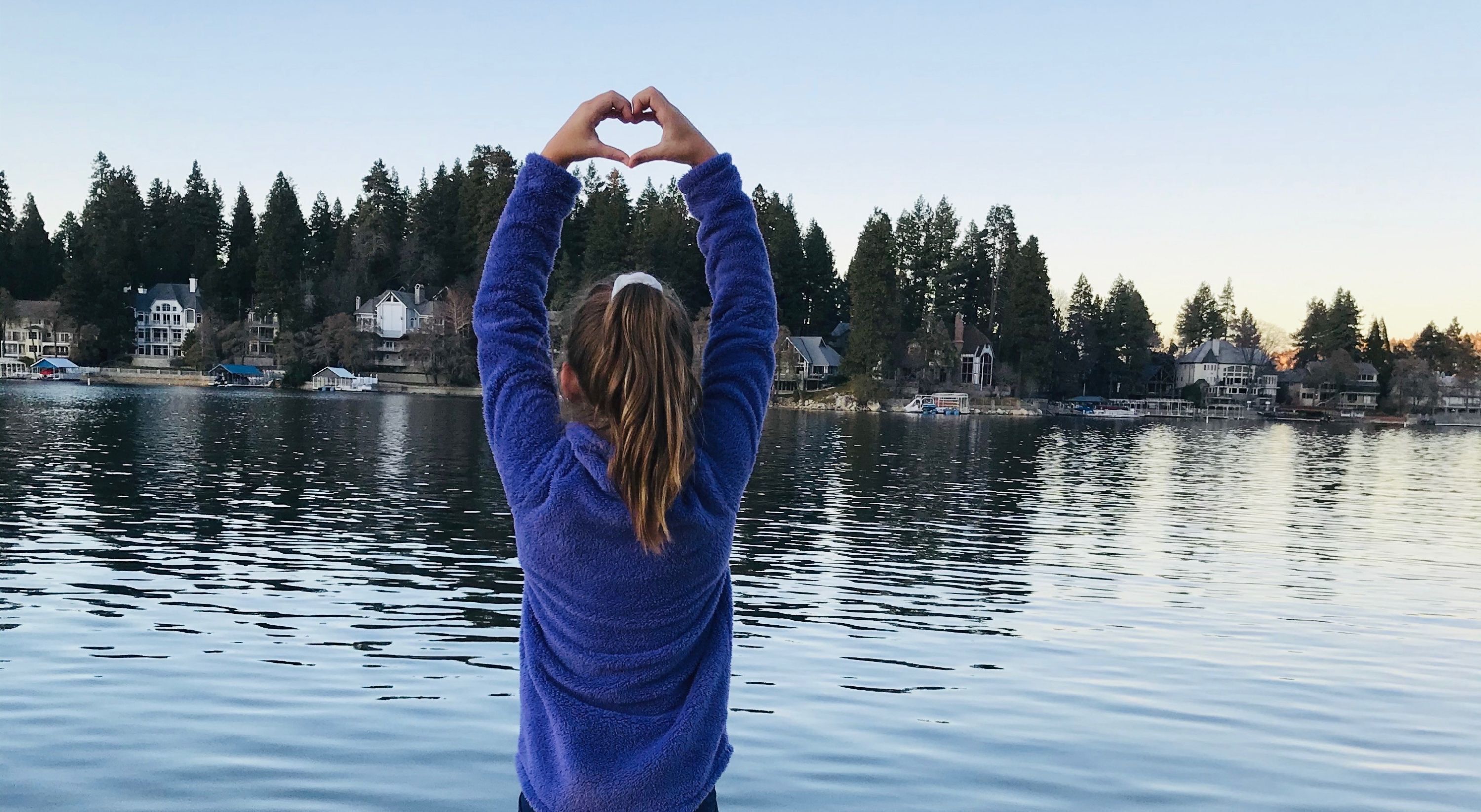 A person making a heart shape with their hands over a body of water with houses in the background.