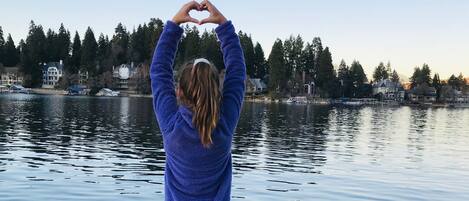 A person making a heart shape with their hands over a body of water with houses in the background.