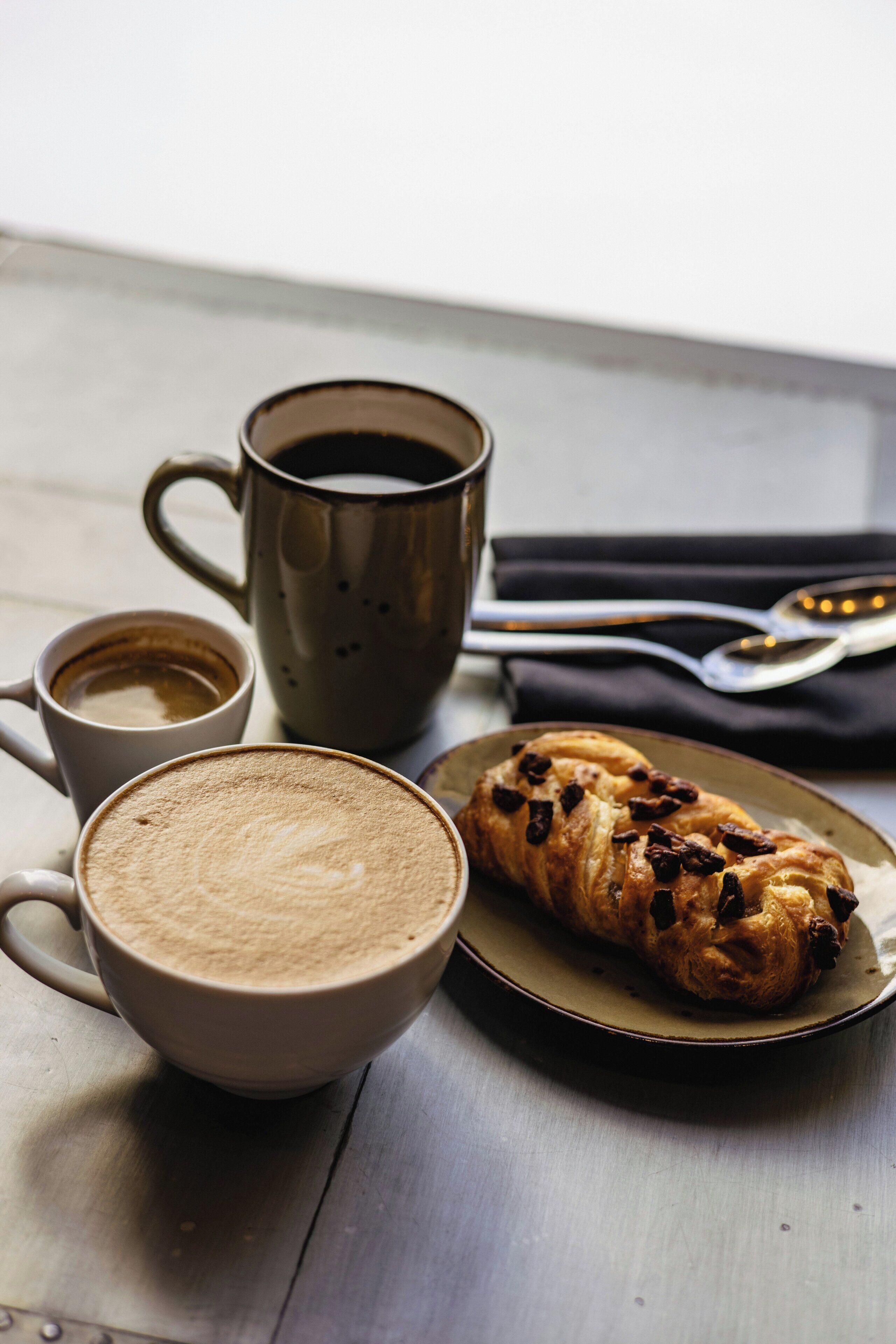 A table with a metal mug, a cup of coffee, and a pastry on a plate.