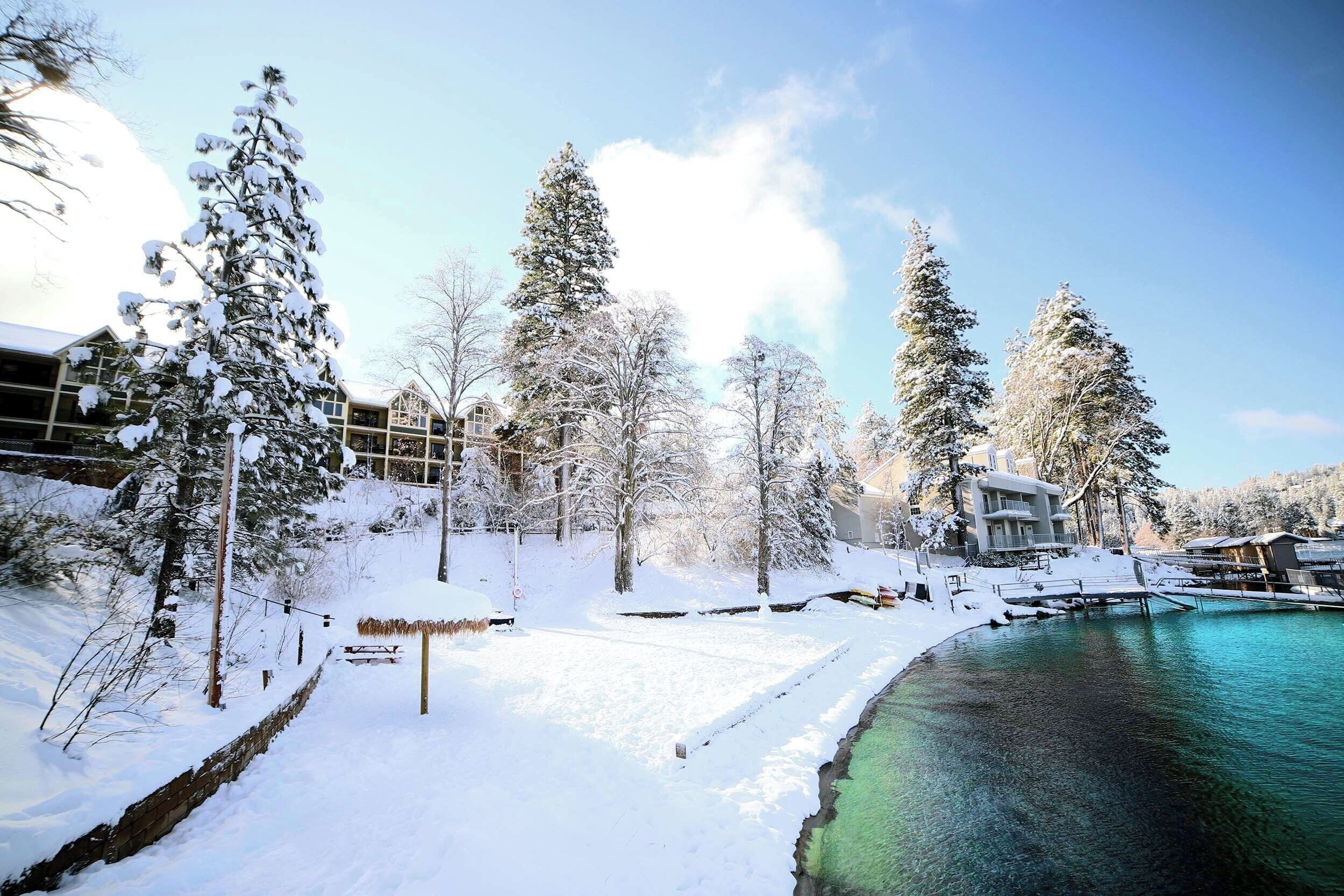 Un paisaje nevado con cielo azul despejado, una masa de agua y varios edificios con nieve en los tejados.