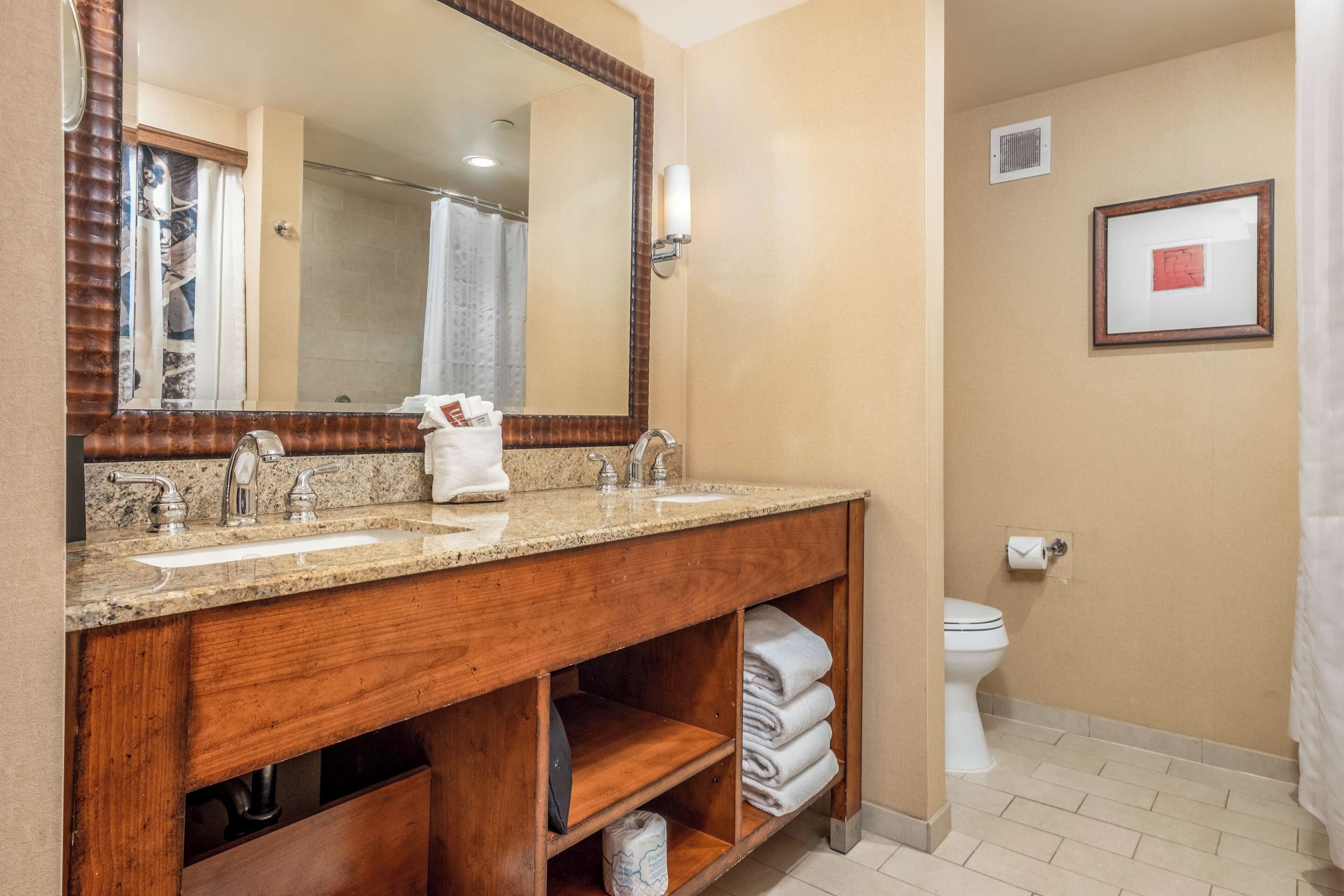A bathroom with a wooden vanity, two sinks, a mirror, a toilet, and a shower curtain.
