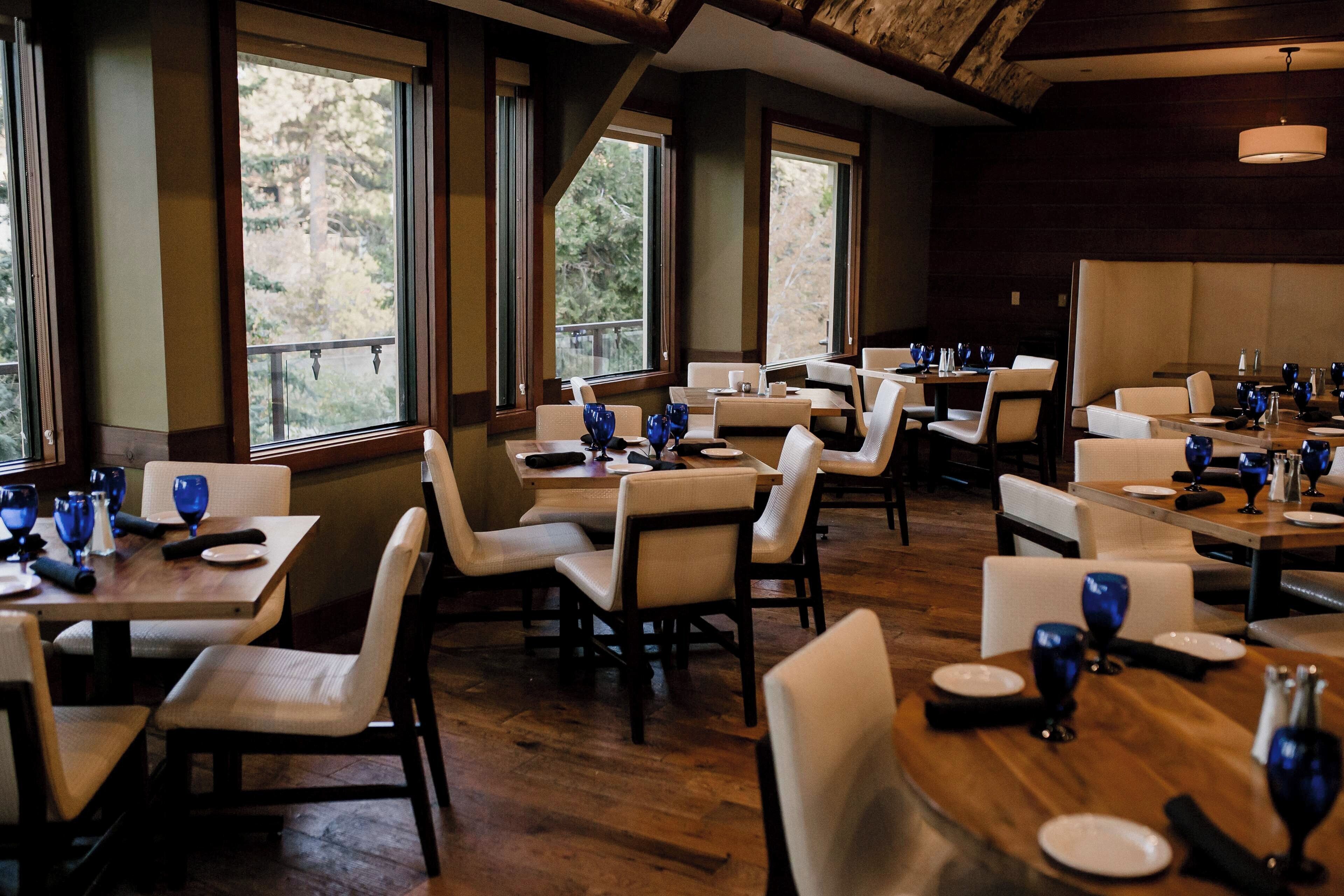 A restaurant with multiple tables and chairs, large windows, and a wooden ceiling.