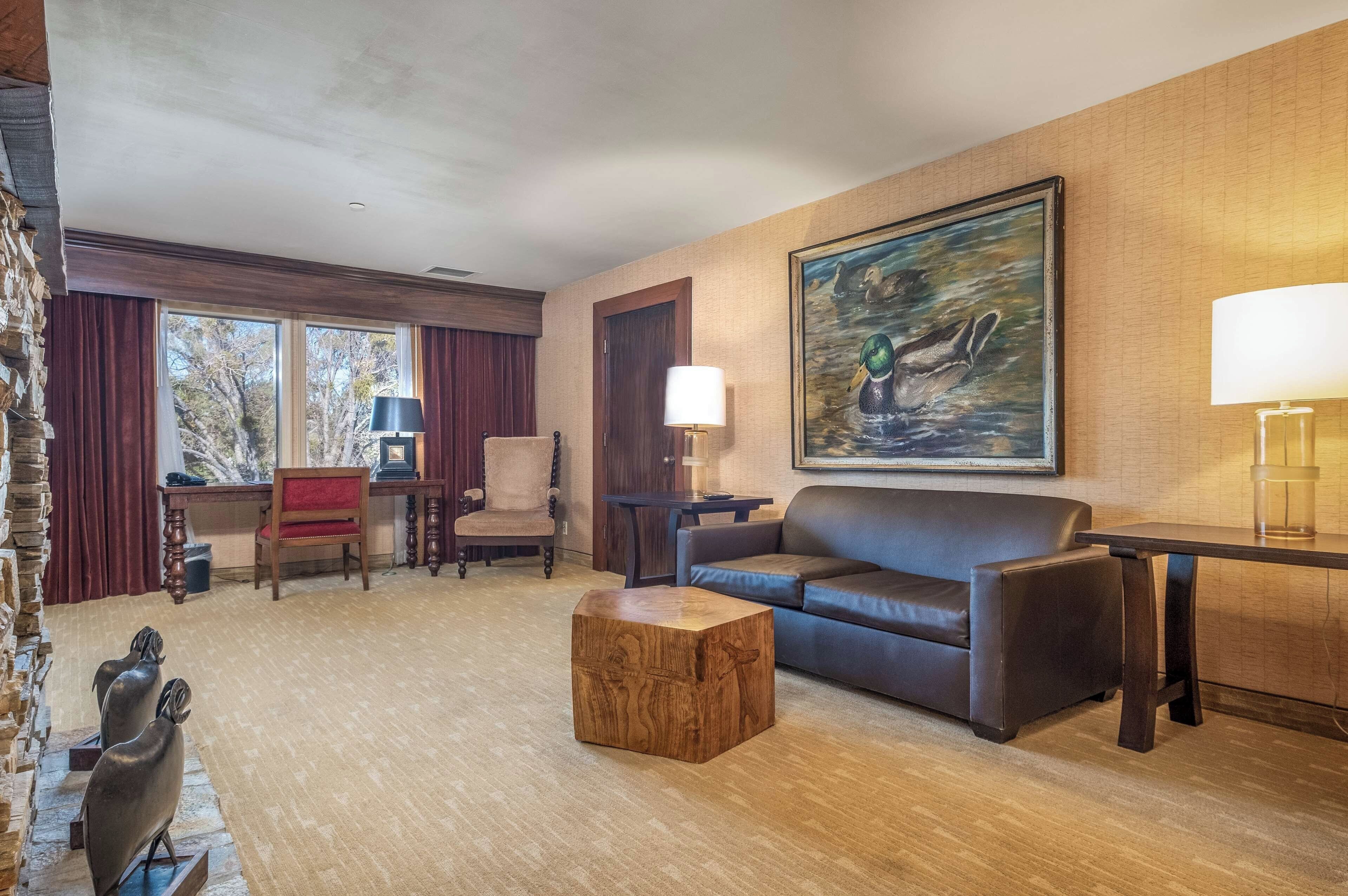 A hotel room with a leather sofa, a wooden coffee table, a painting on the wall, and a desk with a lamp.