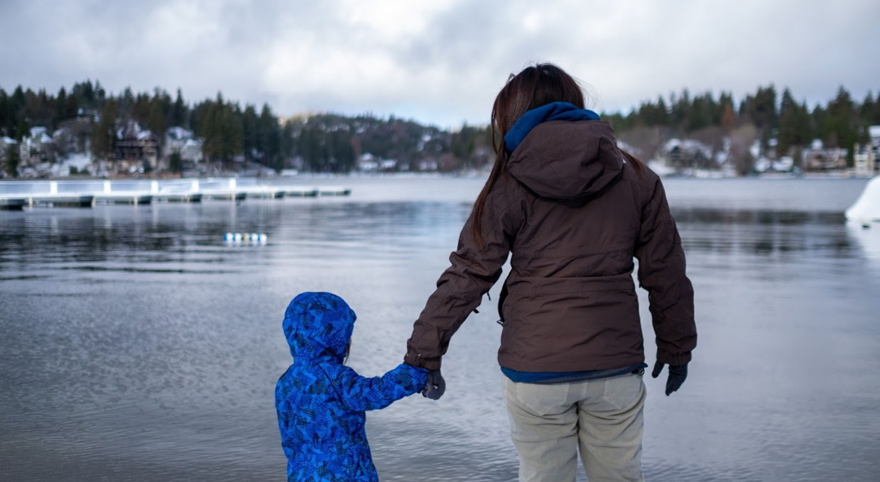 A person in a brown jacket holding hands with a child in a blue jacket, standing by a calm body of water with houses in the background.