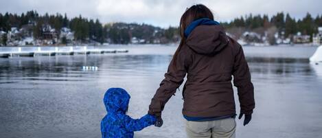 A person in a brown jacket holding hands with a child in a blue jacket, standing by a calm body of water with houses in the background.