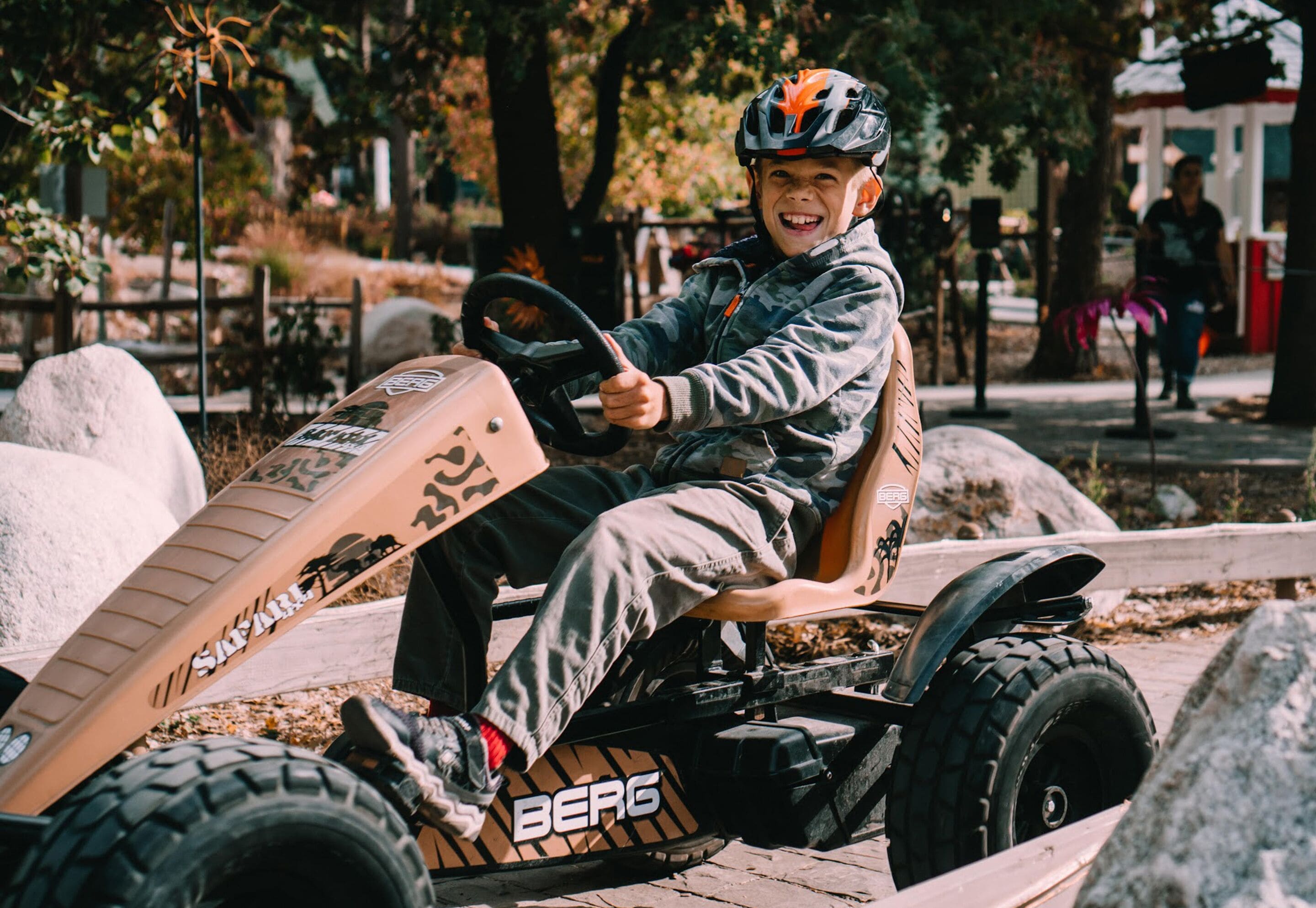 Una persona conduciendo un kart con la marca BERG, en un entorno exterior con árboles y un camino pavimentado.