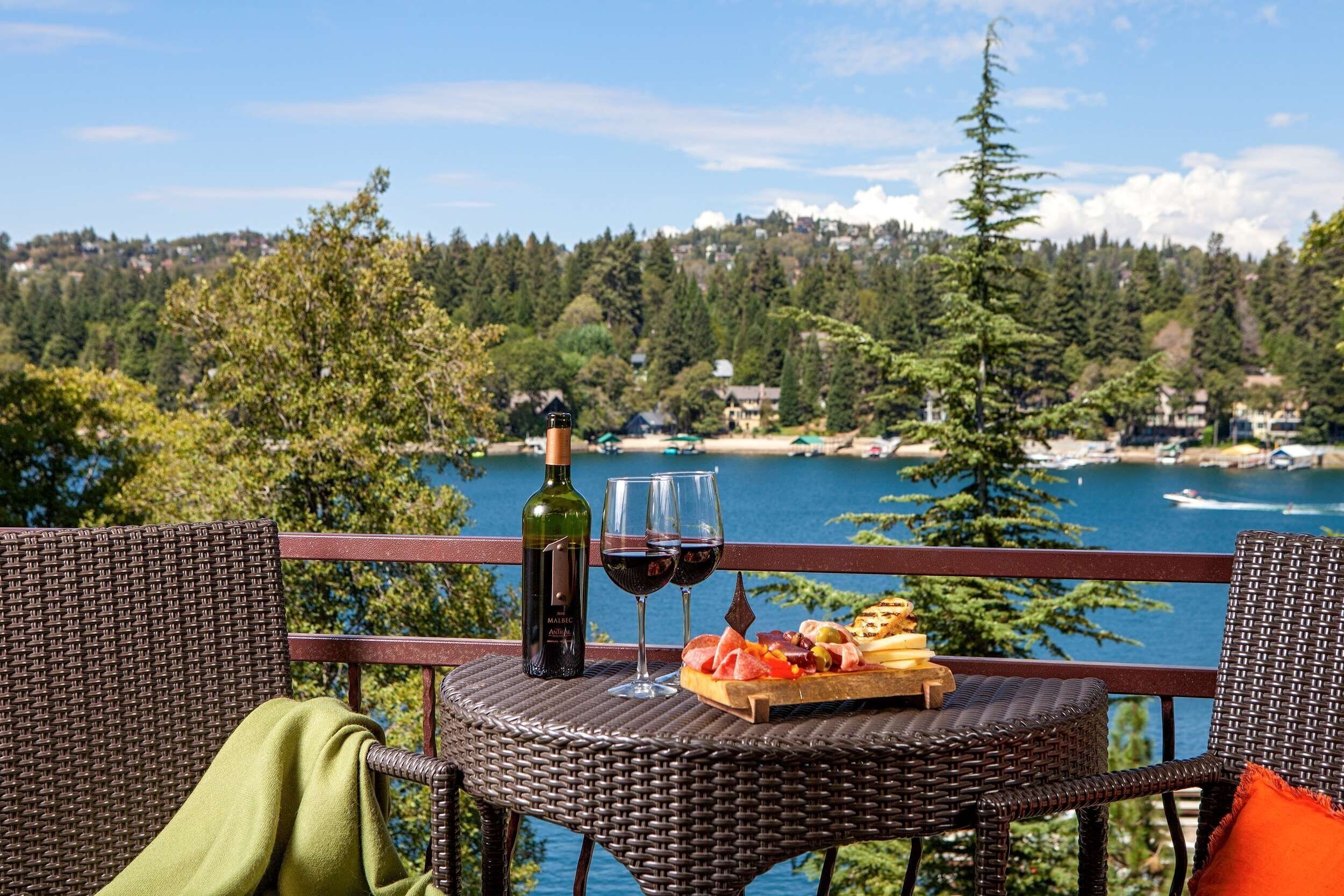 A wicker table with a bottle of wine, glasses, and a charcuterie board overlooking a lake with houses in the background.