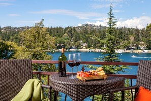 A wicker table with a bottle of wine, glasses, and a charcuterie board overlooking a lake with houses in the background. - Lake Arrowhead Resort and Spa (Lake Arrowhead)