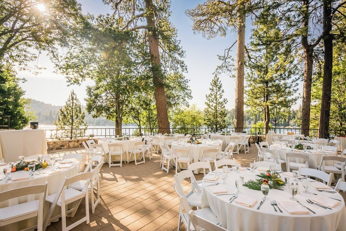 An outdoor dining area with round tables and chairs, set against a backdrop of trees and a body of water.