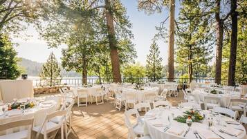 An outdoor dining area with round tables and chairs, set against a backdrop of trees and a body of water.