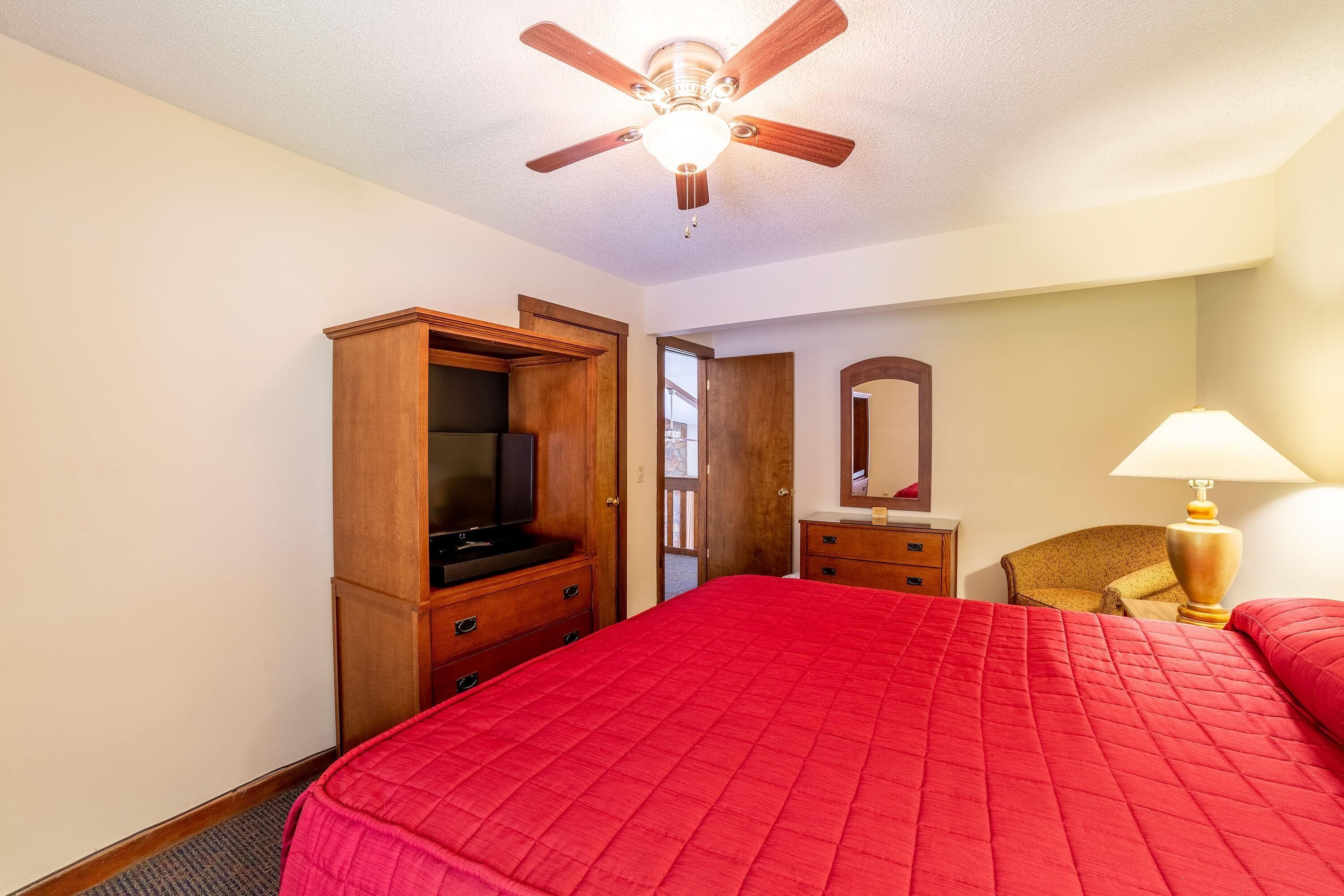 A bedroom with a red bedspread, wooden wardrobe, ceiling fan, and a lamp.