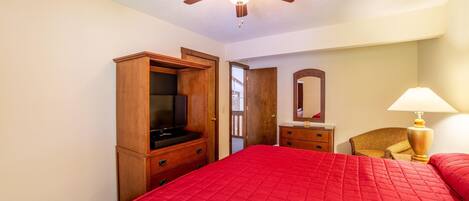 A bedroom with a red bedspread, wooden wardrobe, ceiling fan, and a lamp.