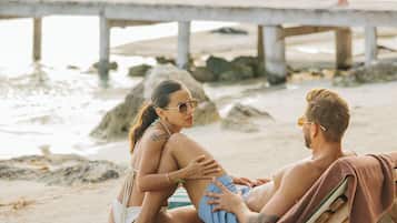 Un couple est assis sur une chaise de plage, une serviette à portée de main. On aperçoit une jetée au loin et une côte rocheuse.