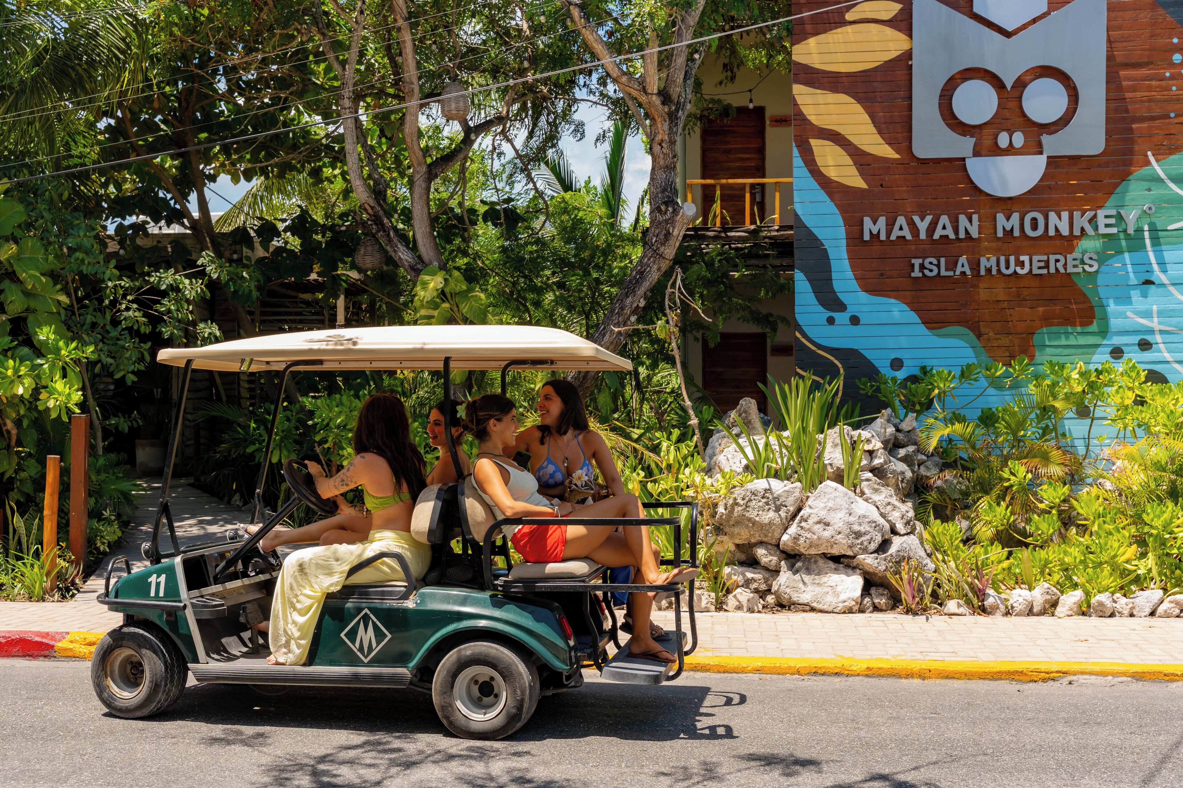 A golf cart with passengers on a street, a mural with a monkey, and tropical vegetation.