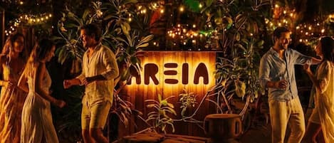 Un entorno tropical al aire libre con un letrero de madera que dice "AREIA", rodeado de plantas y luces de cuerda.