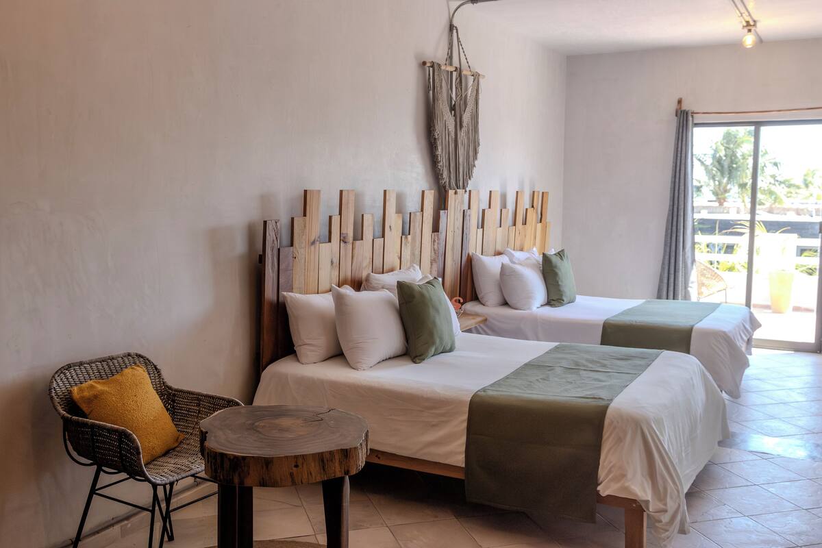 A hotel room with two beds, a wooden headboard, a wicker chair, and a round wooden table.