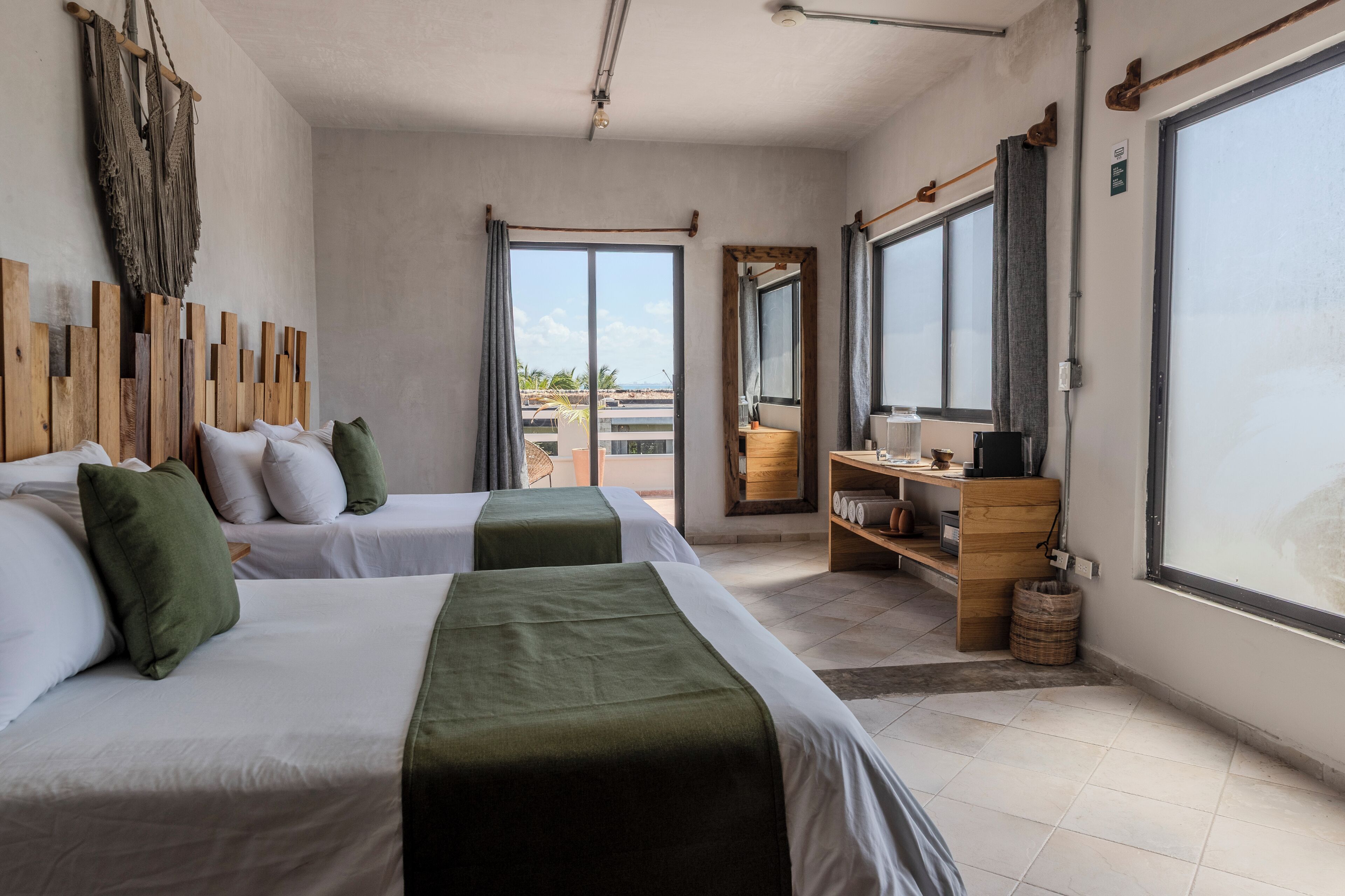 A hotel room with two beds, a wooden shelf with toiletries, a large window, and a view of the outdoors.