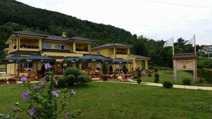 A yellow building with outdoor seating and umbrellas, surrounded by greenery and hills. - Akva Gold Apartmani (Arandelovac)