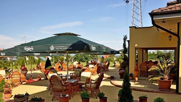 An outdoor seating area with wicker furniture and green umbrellas, surrounded by potted plants and a building with a tiled roof. - Akva Gold Apartmani (Arandelovac)