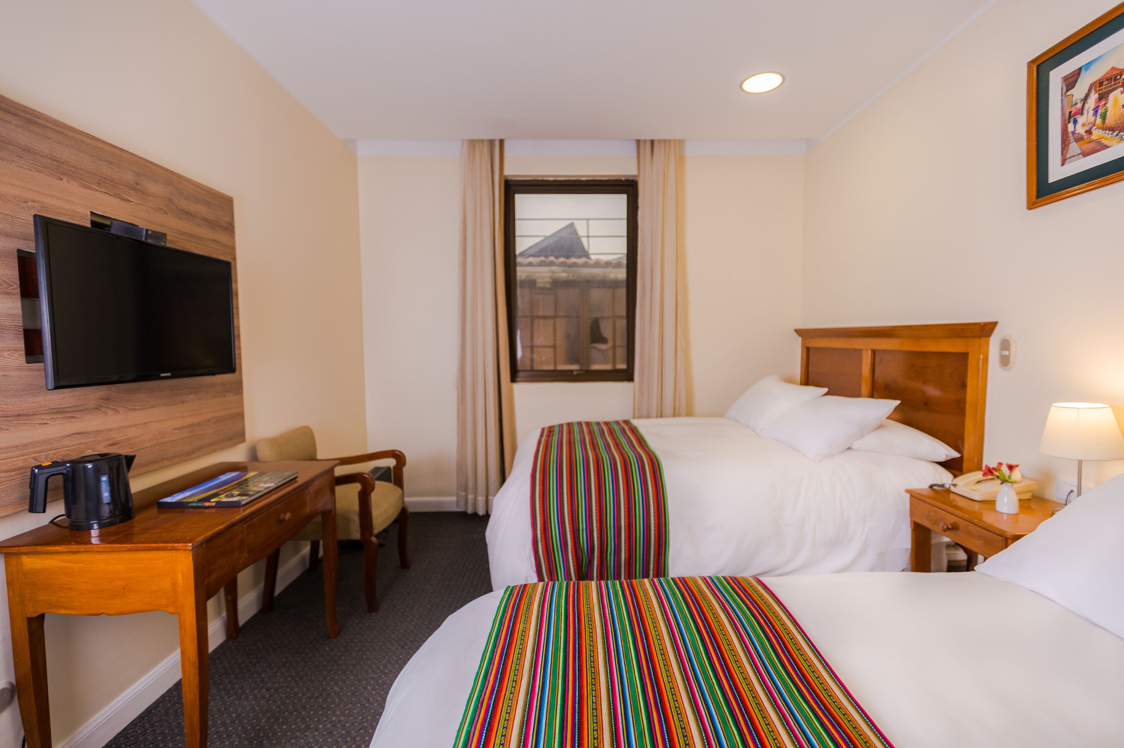 Habitación de hotel con dos camas, un televisor, un escritorio con cafetera y una ventana que ofrece vistas a un edificio.