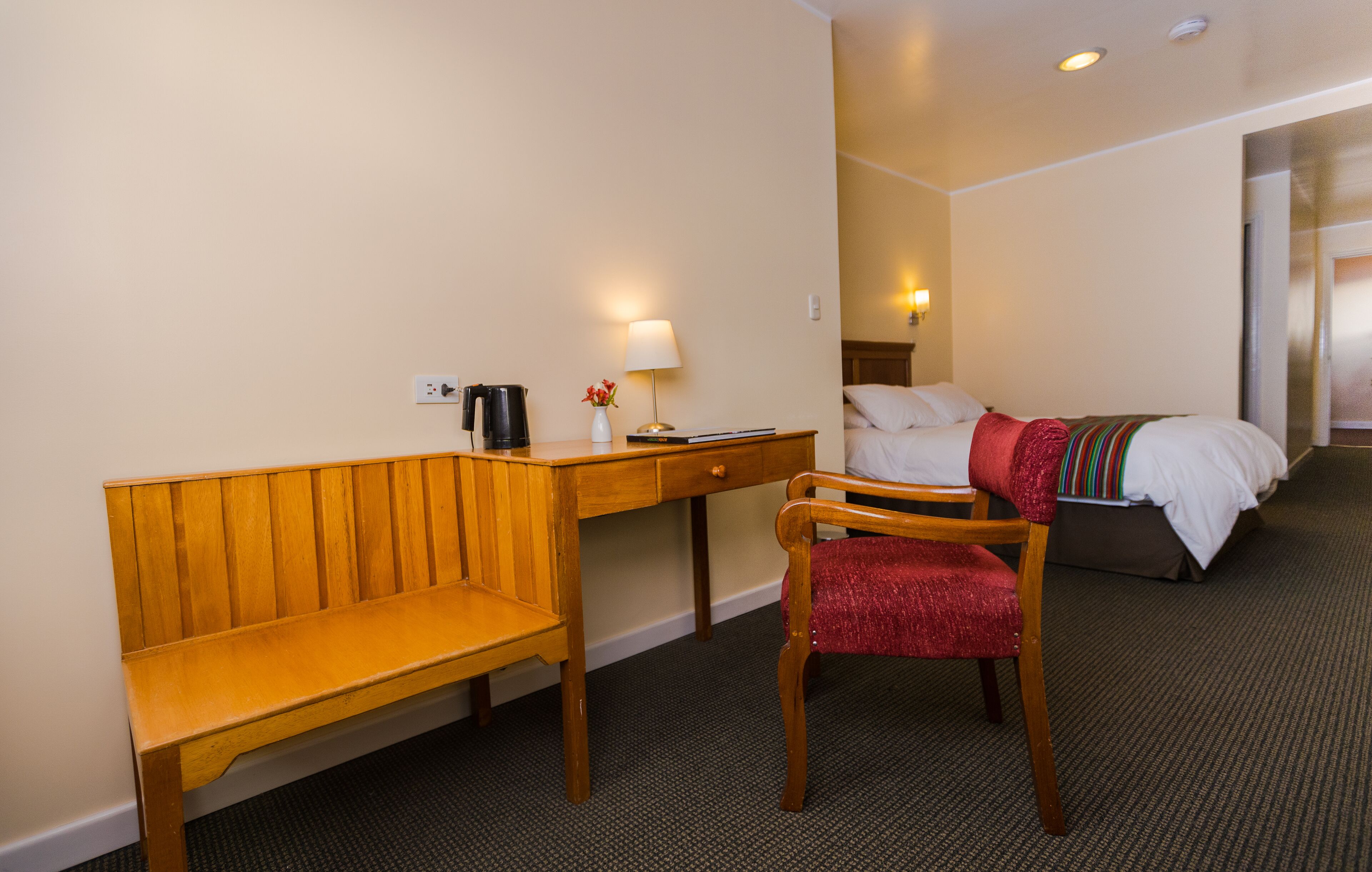 Una habitación de hotel con un escritorio de madera, una silla, una cama, una lámpara y una mesita de noche.