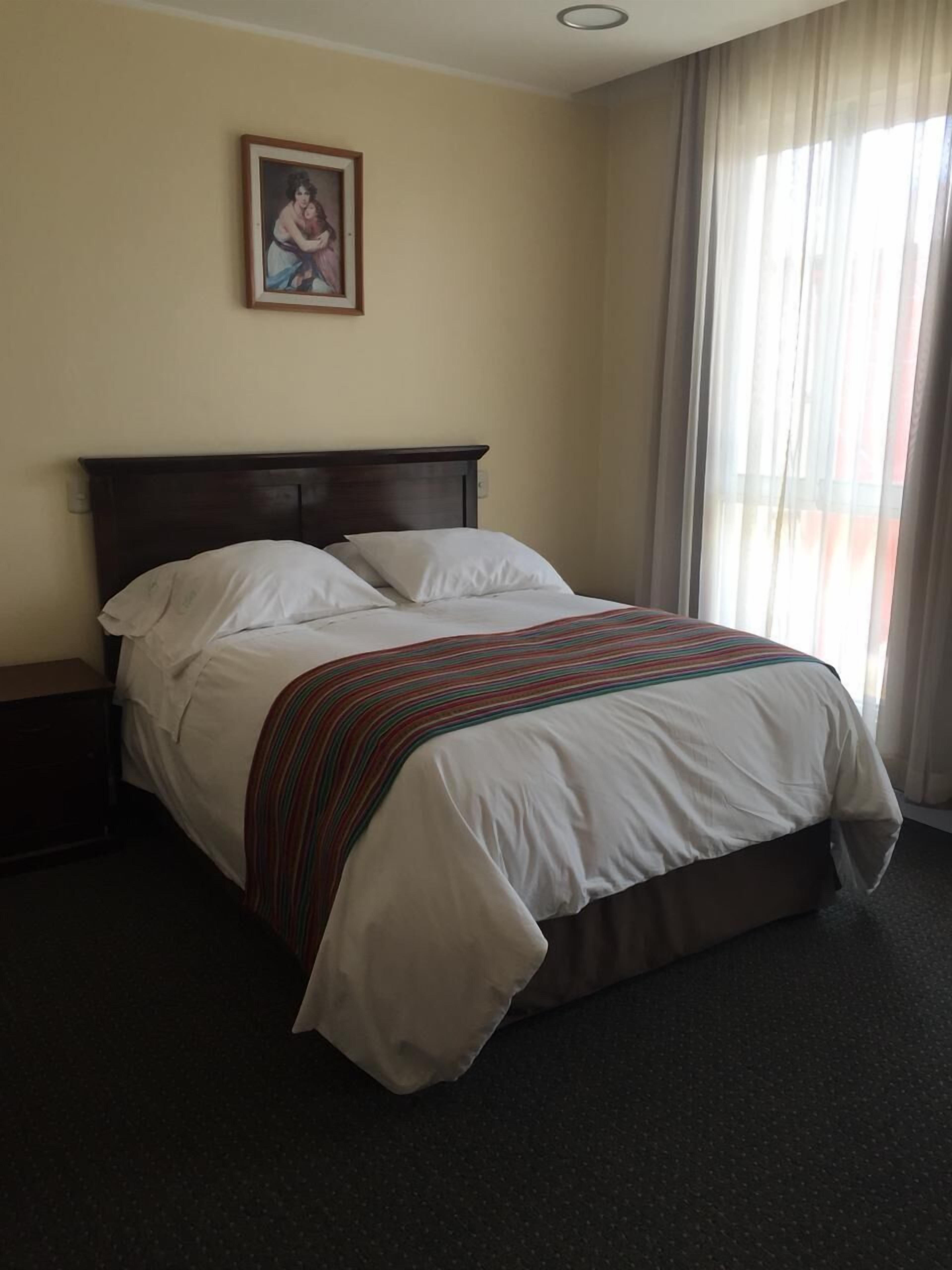 Una cama bien hecha con sábanas blancas y una manta a rayas, cabecera de madera, mesita de noche, un cuadro en la pared y una ventana con cortinas traslúcidas.
