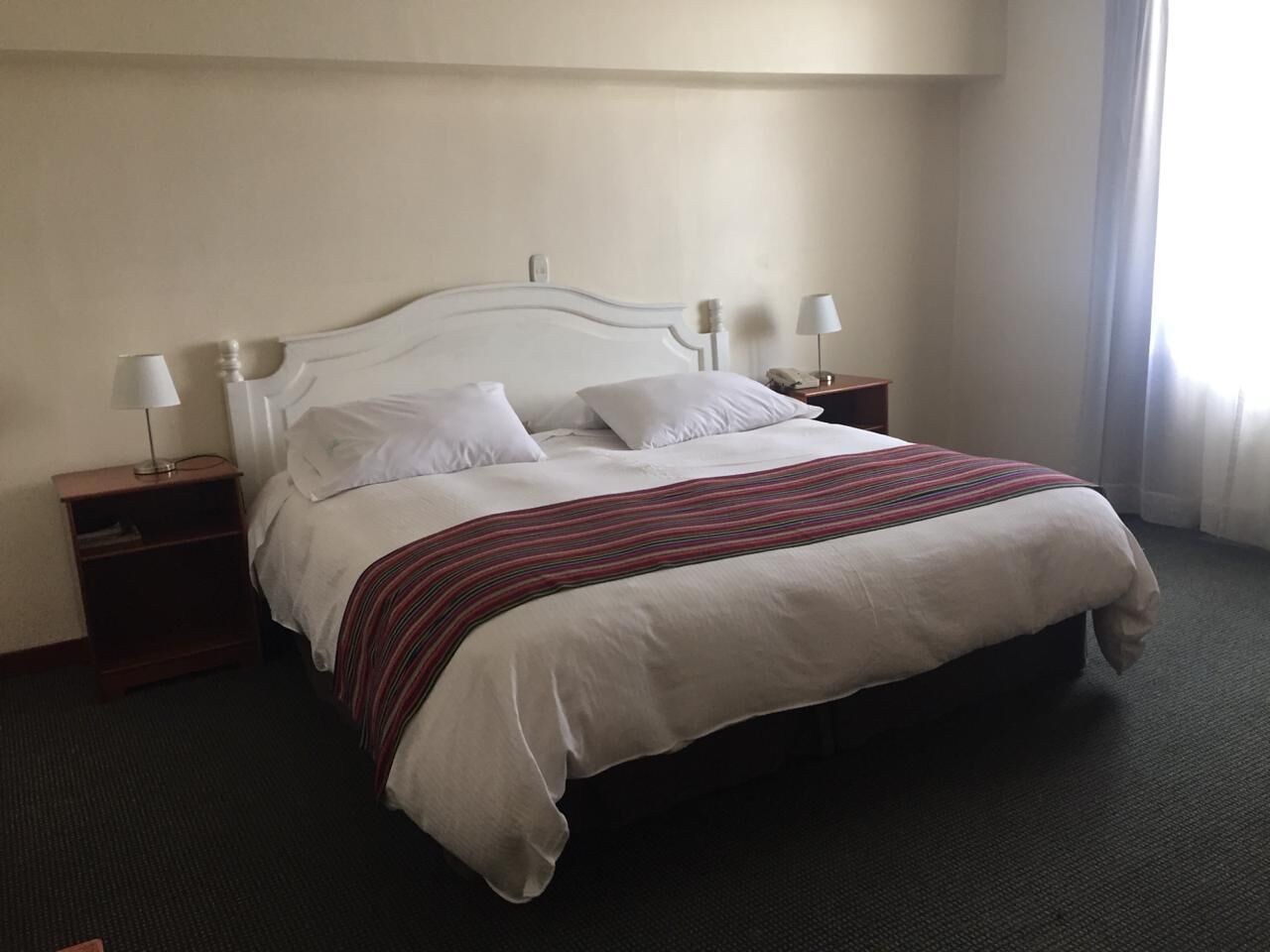 Habitación de hotel con una cama grande, dos mesitas de noche con lámparas, una colcha a rayas y una ventana con cortinas.