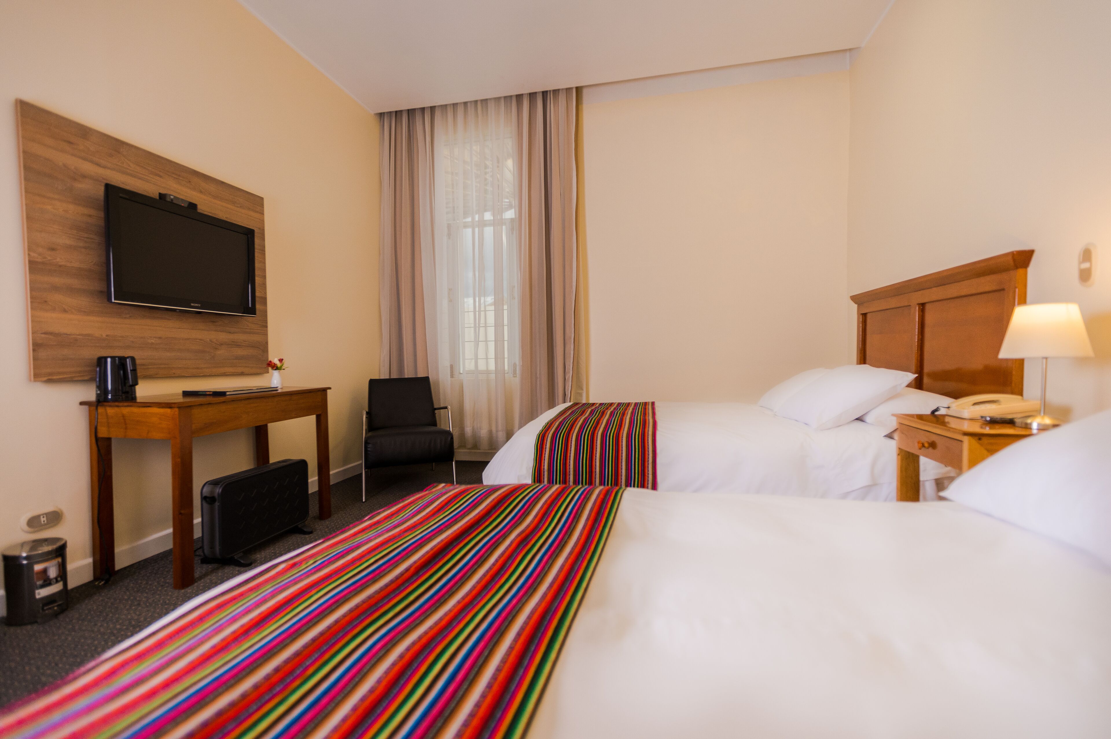 A hotel room with two beds, a TV, a desk with a coffee maker, a chair, and a lamp.
